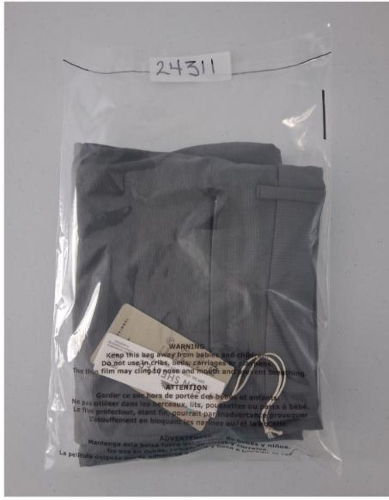K&ouml;nnte beinhalten: Eine gefaltete dunkelgraue Hose in einer durchsichtigen Plastikt&uuml;te. Ein wei&szlig;es Etikett mit der Nummer 24311 ist oben an der T&uuml;te befestigt. Ein beiges Produktetikett mit Warntext ist mit einer wei&szlig;en Schnur an der Hose befestigt.