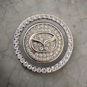 May include: A circular car emblem, featuring the Mazda logo, surrounded by a ring of clear crystals. The emblem has a silver and black color scheme, with a ring of crystals around the outer edge.
