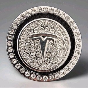 May include: A circular car accessory in silver, with a black edge. The Tesla logo is at the centre, encircled by sparkling crystals. The outer rim is also decorated with crystals, giving it a glamorous appearance.