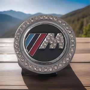 May include: A circular, silver-toned emblem with a black central area displaying the letter "M" adorned with clear crystals. The emblem is edged with clear crystals and features a blue, red, and white stripe design.