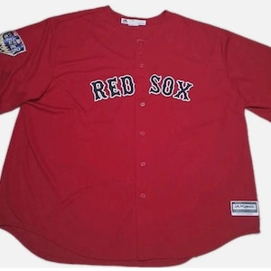 May include: A red baseball jersey featuring the text "RED SOX" in white and black lettering across the chest. The jersey has a button-up front and a patch on the left sleeve. The Majestic brand label is visible.