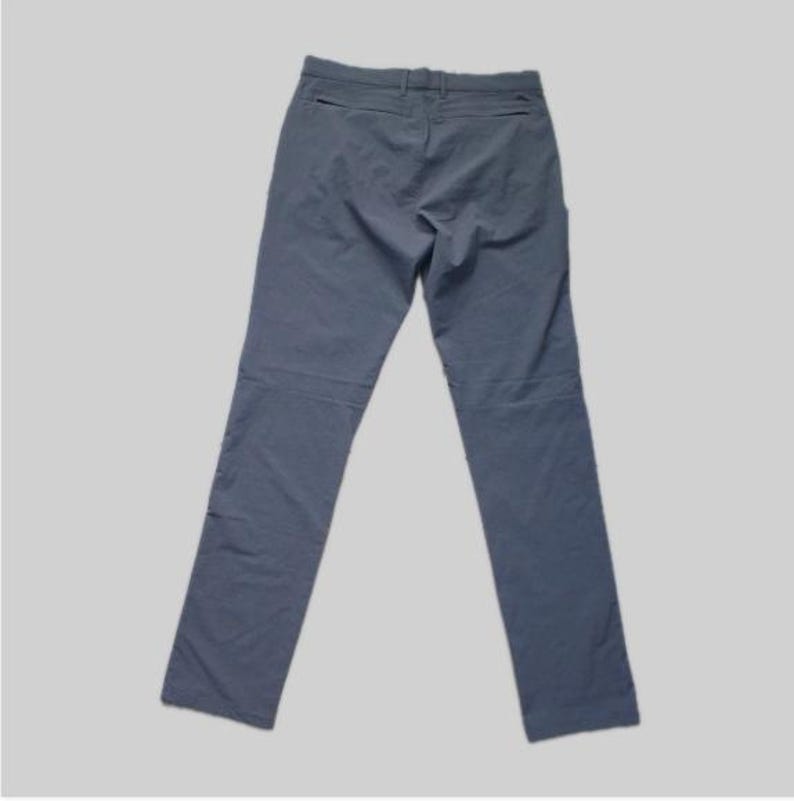 K&ouml;nnte beinhalten: Eine schieferblaue Hose mit einem Bund mit G&uuml;rtelschlaufen, einem Knopfverschluss vorne in der Mitte und zwei horizontalen Rei&szlig;verschlusstaschen direkt unter dem Bund auf der R&uuml;ckseite. Die Hose hat einen geraden Schnitt und eine glatte Oberfl&auml;che.