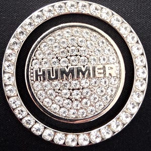 May include: A silver-coloured circular emblem featuring the word "HUMMER" in raised lettering. The emblem is adorned with clear, sparkling rhinestones, with a ring of rhinestones around the outer edge.