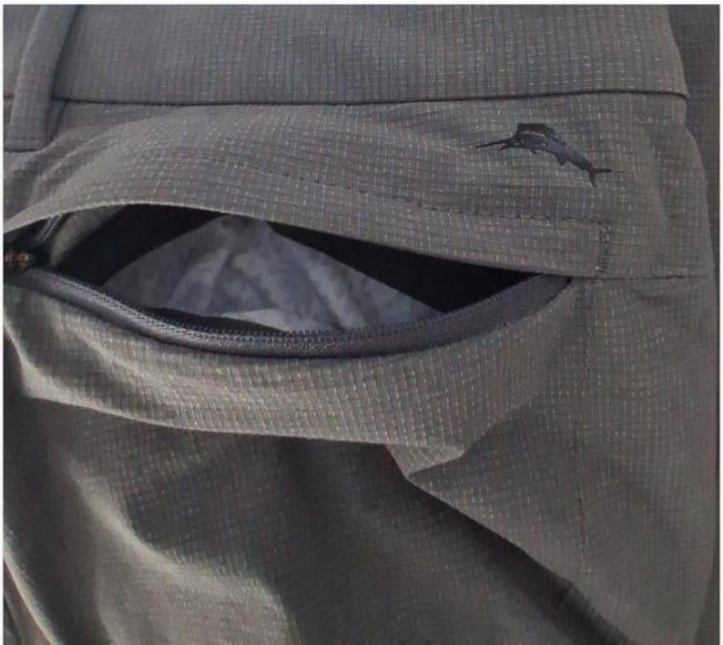 K&ouml;nnte beinhalten: Dunkelolivgr&uuml;ne Hose mit strukturiertem Gittermuster und einer Rei&szlig;verschlusstasche. Ein kleines, schwarz gesticktes Marlin-Fisch-Logo befindet sich &uuml;ber der Taschen&ouml;ffnung, die einen Blick auf ein helles Innenfutter freigibt.