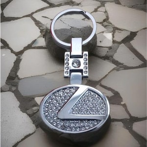 Op de afbeelding: Een zilverkleurige metalen sleutelhanger met een rond embleem met een gestileerde letter L, ingelegd met talloze heldere strass-steentjes. De sleutelhanger bevat een rechthoekig verbindingsstuk dat ook met strass-steentjes is versierd en een ring.