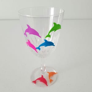 May include: A clear glass wine glass with five colorful dolphin stickers on the side and bottom. The dolphins are pink, green, blue, orange, and red.