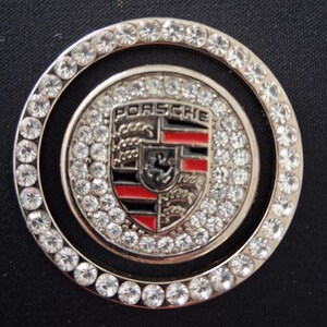 May include: A circular silver emblem with the Porsche logo at its centre, encircled by a ring of clear rhinestones. The Porsche crest features red and black detailing. The emblem is designed to be decorative.