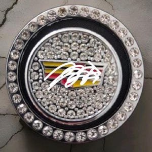 May include: A circular Cadillac emblem badge featuring a silver-tone metallic frame encrusted with clear rhinestones. The centre displays the Cadillac crest in yellow, red, and black, surrounded by a dense field of clear rhinestones.