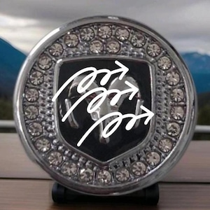 May include: A circular chrome emblem featuring a central black shield with a silver ram head logo. The outer border is encrusted with clear, sparkling rhinestones. Three white, hand-drawn arrow graphics are overlaid across the centre of the emblem.