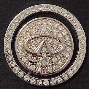 Op de afbeelding: Een ronde, zilverkleurige embleem met het Infiniti-logo in het midden, ingelegd met talrijke heldere, sprankelende strass-steentjes. Het embleem is omgeven door een ring van kleinere, eveneens versierde strass-steentjes. De achtergrond is zwart.