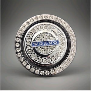 May include: A circular car emblem in chrome, featuring the word "VOLVO" in blue. The emblem is embellished with clear rhinestones around the edge and in the centre.