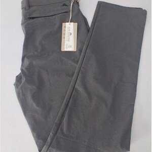 K&ouml;nnte beinhalten: Eine einfarbige graue Hose mit einer Rei&szlig;verschlusstasche unter dem Bund, G&uuml;rtelschlaufen und einem rechteckigen beigen Produktanh&auml;nger, der mit einer Schnur befestigt ist. Das Etikett zeigt den Text SUN SHELTER sowie kleine Symbole und Details.