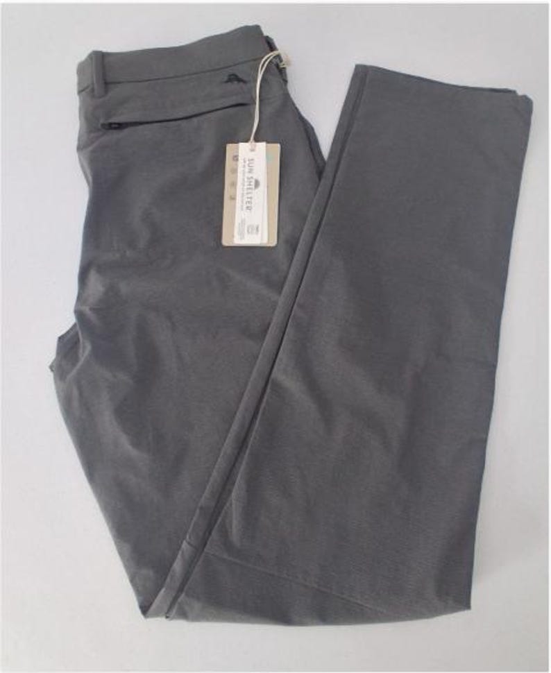 K&ouml;nnte beinhalten: Eine einfarbige graue Hose mit einer Rei&szlig;verschlusstasche unter dem Bund, G&uuml;rtelschlaufen und einem rechteckigen beigen Produktanh&auml;nger, der mit einer Schnur befestigt ist. Das Etikett zeigt den Text SUN SHELTER sowie kleine Symbole und Details.