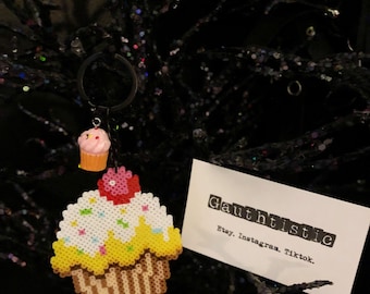 May include: A beaded cupcake keychain with yellow icing, white sprinkles, and a pink cherry on top. The keychain is attached to a small, pink cupcake charm. The keychain is hanging from a black metal ring. The background is a black, sparkly fabric.