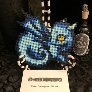 May include: A blue and white pixel art character with a yellow eye, made with perler beads. The character is displayed on a black and white stand. The image also includes a small bottle with a "Death Potion" label.