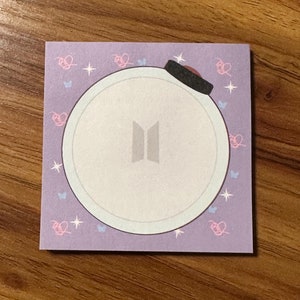 May include: Purple notepad with a white circle containing the BTS logo and a black hat on top. The circle is surrounded by pink hearts and blue butterflies.