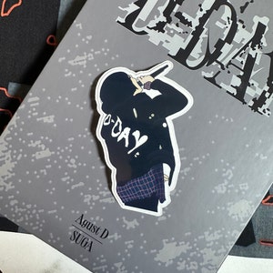 May include: A gray album cover with the word "D-DAY" printed in black and white. The album cover features a sticker of a person wearing a black jacket and a plaid shirt, holding a microphone. The text "Agust D Suga" is printed in black on the album cover.