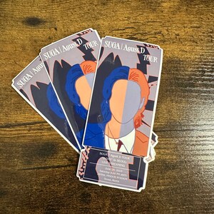 May include: Three colorful stickers with a stylized illustration of a person's face. The stickers are for the "SUGA | Agust D TOUR" and include the text "D-DAY in SEOUL" and "LIVE STREAMING". The date and time of the event are June 25, 2023 at 5:00 PM (KST) at the JAMSIL INDOOR STADIUM in Seoul, Korea.