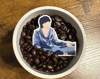 Pode incluir: Uma tigela de cerâmica branca cheia de grãos de café castanho escuro. Um adesivo de um personagem de desenho animado vestindo uma jaqueta azul é colocado em cima dos grãos.