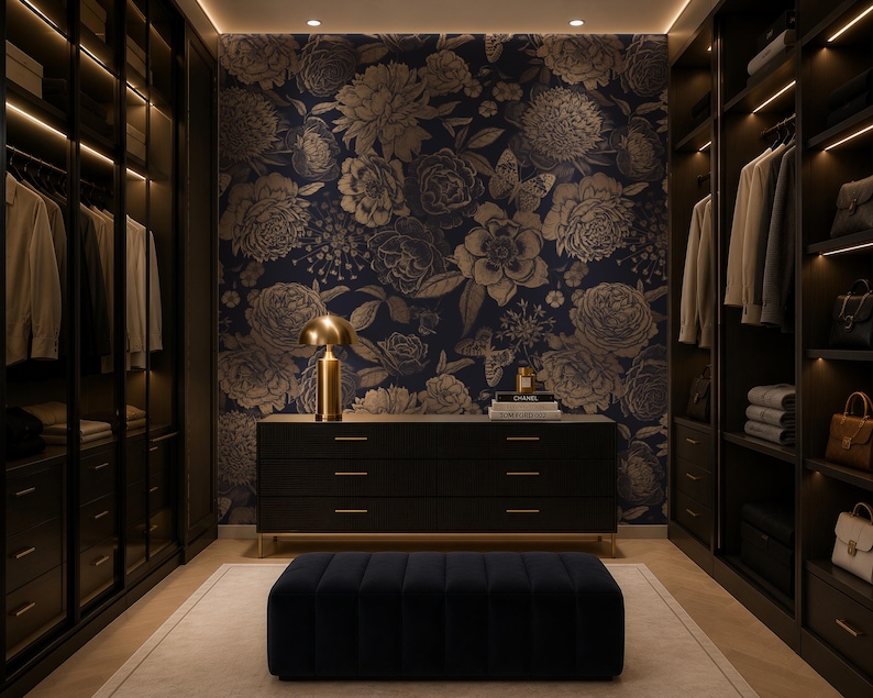 May include: A walk-in closet featuring dark wood cabinetry with integrated lighting, a central black dresser with gold hardware, a navy blue velvet ottoman, and a dark blue wallpaper backdrop with a gold floral and butterfly pattern.