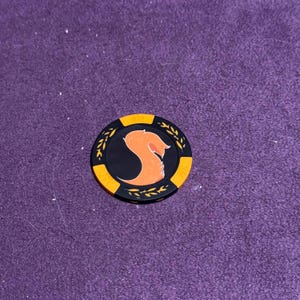 Puede incluir: Una ficha de p&oacute;quer negra y naranja con un dise&ntilde;o estilizado de cabeza de zorro naranja. La ficha tiene un centro negro con la cabeza de zorro y un borde dorado con detalles de hojas. La ficha est&aacute; sobre una superficie morada.