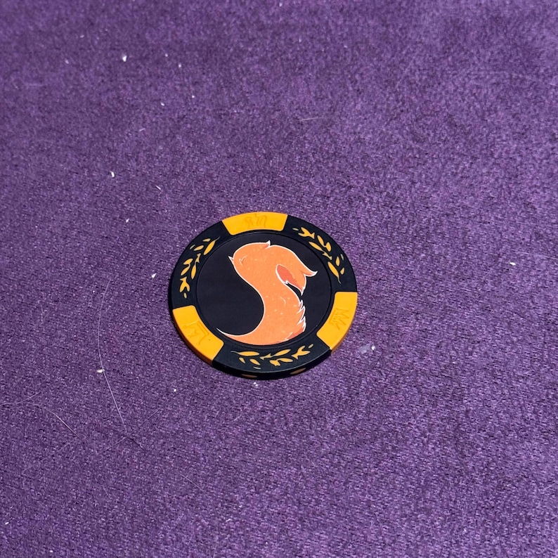 Puede incluir: Una ficha de p&oacute;quer negra y naranja con un dise&ntilde;o estilizado de cabeza de zorro naranja. La ficha tiene un centro negro con la cabeza de zorro y un borde dorado con detalles de hojas. La ficha est&aacute; sobre una superficie morada.