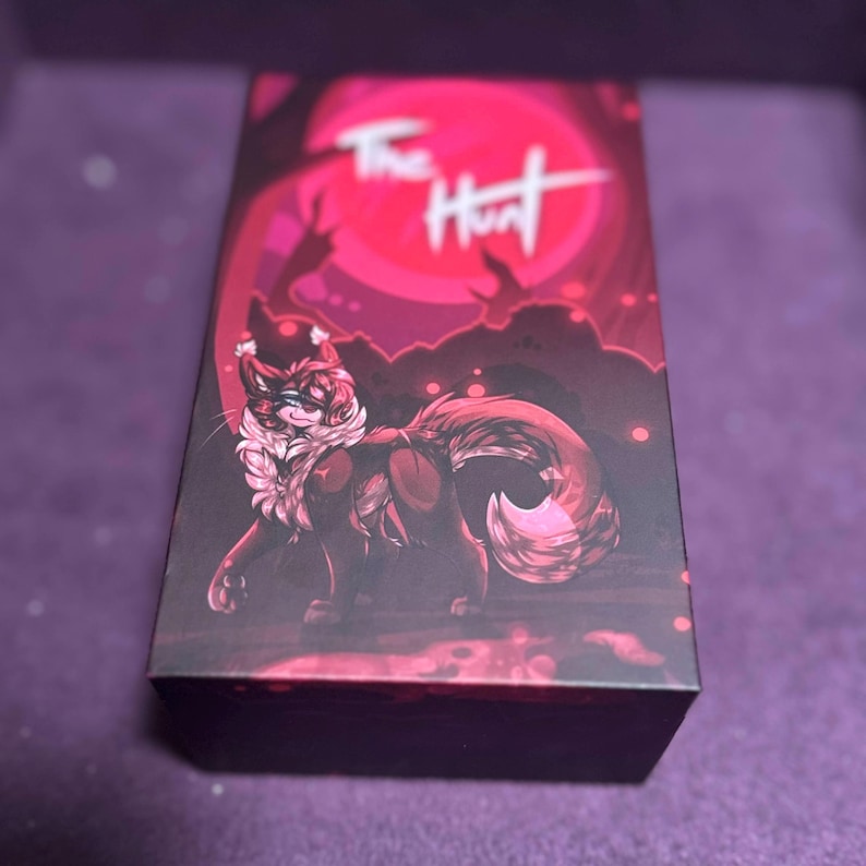 Puede incluir: Una caja oscura con una ilustraci&oacute;n de fantas&iacute;a de una criatura parecida a un zorro con una melena y cola blancas y esponjosas. La caja tiene las palabras "The Hunt" en escritura blanca sobre un fondo rojo con una luna llena y siluetas de &aacute;rboles.
