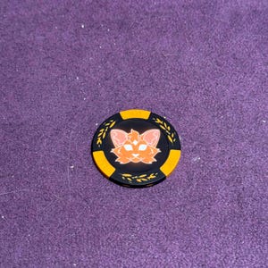 Puede incluir: Ficha de p&oacute;quer negra y naranja con un dise&ntilde;o de cara de gato. La ficha tiene un borde amarillo y detalles de hojas. La cara del gato es naranja con orejas rosas. Aproximadamente 4 cm de di&aacute;metro.