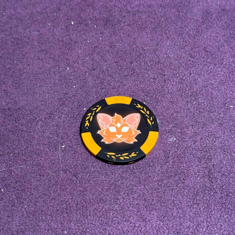 Puede incluir: Ficha de p&oacute;quer negra y naranja con un dise&ntilde;o de cara de gato. La ficha tiene un borde amarillo y detalles de hojas. La cara del gato es naranja con orejas rosas. Aproximadamente 4 cm de di&aacute;metro.