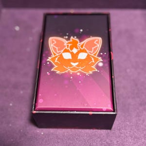Puede incluir: Caja rectangular con un dise&ntilde;o degradado de rosa y morado. La parte superior de la caja presenta una ilustraci&oacute;n de cara de gato naranja con un contorno brillante. La caja est&aacute; sobre un fondo morado.