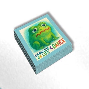 May include: A stack of square stickers with a cartoon frog illustration. The frog is green with a red zig-zag mouth and sad eyes. Text on the sticker reads "DARKSIDE OF LIFE < DANCE" in colorful letters. The stickers have a light blue border.