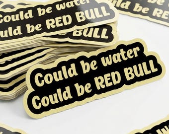 May include: A stack of stickers with a black background and a cream-colored border. The text "Could be water, could be RED BULL" is printed in cream-colored font. Several stickers are scattered around the stack.