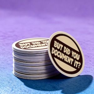 May include: A stack of oval stickers with the text "BUT DID YOU DOCUMENT IT?" in a brown and white design. The stickers are stacked on a blue and purple surface.