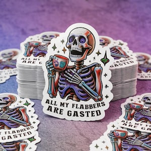 May include: A stack of white stickers featuring a colorful skeleton holding a teacup. The skeleton is depicted with red, purple, and blue hues. The text "ALL MY FLABBERS ARE GASTED" is printed on the sticker in black.