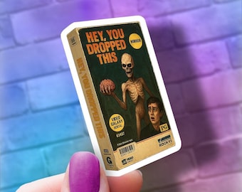 May include: A sticker featuring a vintage VHS tape design with the text "HEY, YOU DROPPED THIS." The artwork depicts a skeletal figure holding a brain, with a terrified person. The tape is labeled "HORROR" and "VIDEO GALAXY RENTAL."