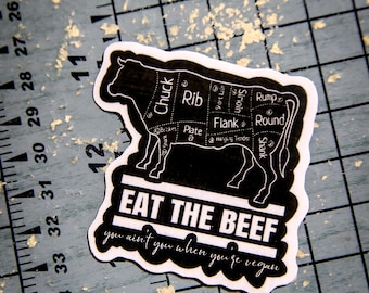 May include: A black and white sticker shaped like a cow with a butcher's cut diagram. The text reads EAT THE BEEF and you ain't you when you're vegan. A ruler is visible on the left showing measurements in inches.
