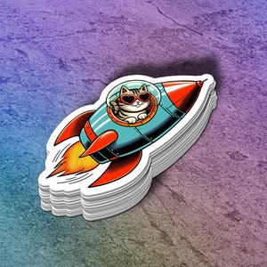 May include: A stack of stickers featuring a cartoon cat in a rocket ship. The cat wears heart-shaped sunglasses and waves. The rocket is teal, red, and orange with a yellow flame.