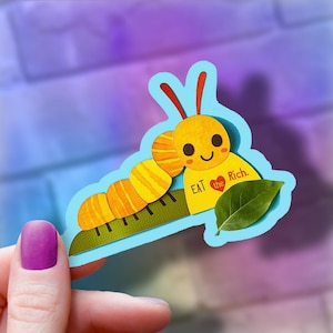 May include: A die-cut sticker featuring a cartoon caterpillar with a yellow body and orange stripes. The caterpillar has a smiling face and the text "EAT the Rich" on its body. A green leaf is also included in the design.