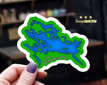 May include: A green and blue sticker with an outline of Lake Winnipesaukee. The sticker has a white border and the text "Lake Winnipesaukee" is written in white.