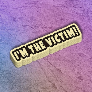 May include: A stack of stickers with the text "I'M THE VICTIM!" in bold, black letters. The text is outlined in black and set against a cream-colored background. The stickers are on a purple and blue textured surface.