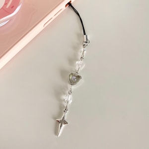 May include: A silver phone charm with a heart pendant, a star charm, and clear crystal beads. The charm is attached to a black string.
