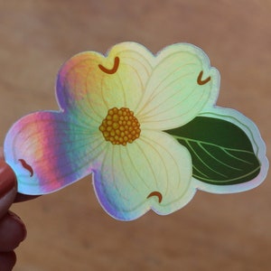 May include: A holographic sticker of a white flower with a yellow center and a green leaf. The flower has a rainbow iridescent finish.