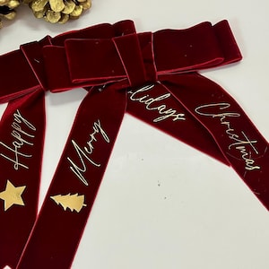 Puede incluir: Lazos navideños de terciopelo burdeos con texto dorado que dice "Happy", "Merry" y "Christmas". Cada lazo tiene una estrella dorada o un adorno de árbol de Navidad. Decoración festiva navideña.