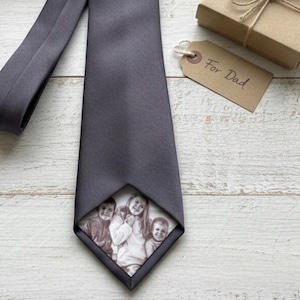 Pode incluir: Uma gravata cinza carvão com uma impressão fotográfica de um retrato de família na ponta. Exibida sobre uma superfície de madeira clara, ao lado de uma caixa de presente e uma etiqueta com "For Dad".