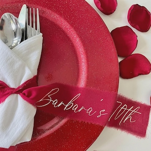 May include: A red plate with a white napkin, silverware, and a red ribbon with the text "Barbara's 70th". Scattered around the plate are red rose petals. The setting is festive and celebratory.