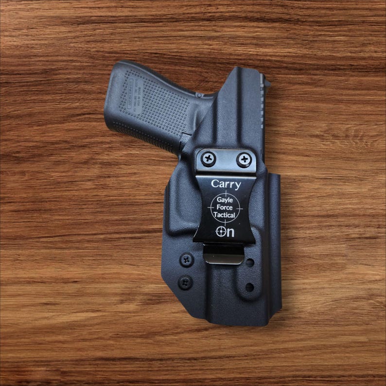 K&ouml;nnte beinhalten: Eine schwarze Holster mit einer Handfeuerwaffe darin, auf einer Holzoberfl&auml;che. Die Holster hat einen Metallclip mit den Worten "Carry", "Gayle Force Tactical" und "On" aufgedruckt. Die Handfeuerwaffe ist schwarz.