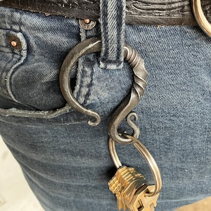 May include: A black metal key hook with a twisted design, attached to a silver key ring with a set of keys. The hook is attached to a pair of blue jeans.