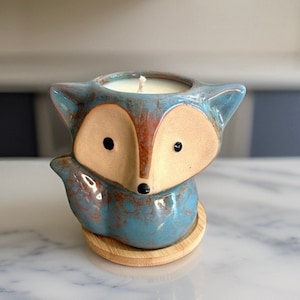 May include: A blue and brown ceramic fox candle holder with a white candle inside. The holder has a wooden base.