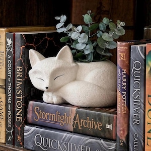 May include: A cream-colored, textured fox-shaped planter with a sleeping expression, featuring pointed ears and a curled tail. The planter holds a small green eucalyptus plant and is positioned on top of a stack of hardcover books.
