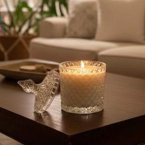 May include: A lit candle in a clear glass jar with a diamond pattern, and a glass lid with a decorative handle. The candle sits on a dark brown wooden table. The background includes a sofa and plants.